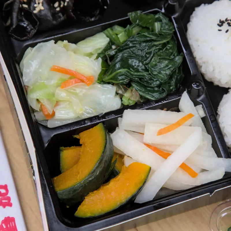 紅蕃茄客製化便當中的配菜近照，包含滷豆干、爽脆的醃蘿蔔、清甜的烤南瓜，每一樣配菜都經過精心處理，色彩繽紛，讓便當更加豐富且健康。