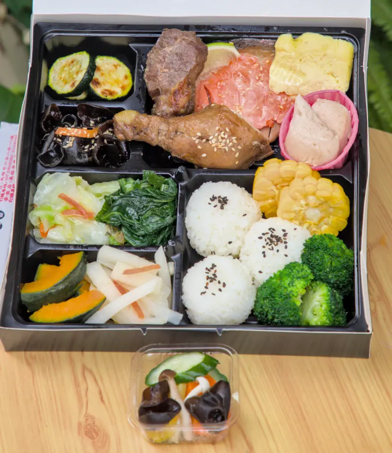 紅蕃茄客製化便當的俯視圖，便當盒分格擺放著多樣主菜與配菜，如滷雞腿、生食級鮭魚、烤櫛瓜、黑木耳等，色彩繽紛，看起來既美味又健康。