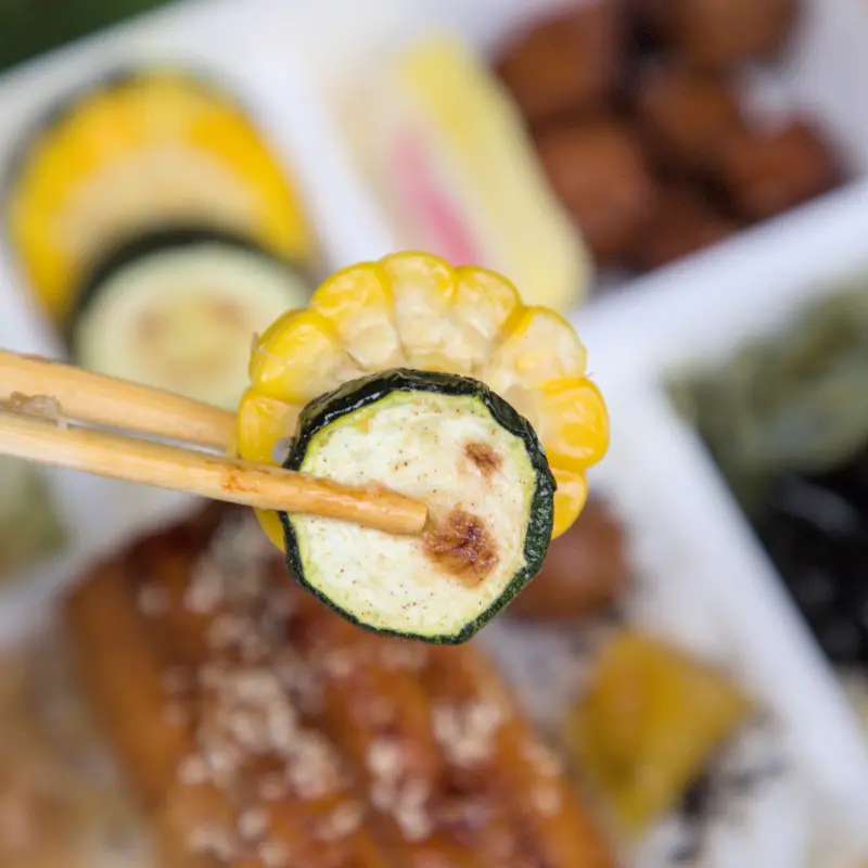 紅蕃茄鰻魚飯的配菜特寫，包含烤玉米與烤櫛瓜，展現便當的多元配菜組合，確保營養均衡，口感豐富。