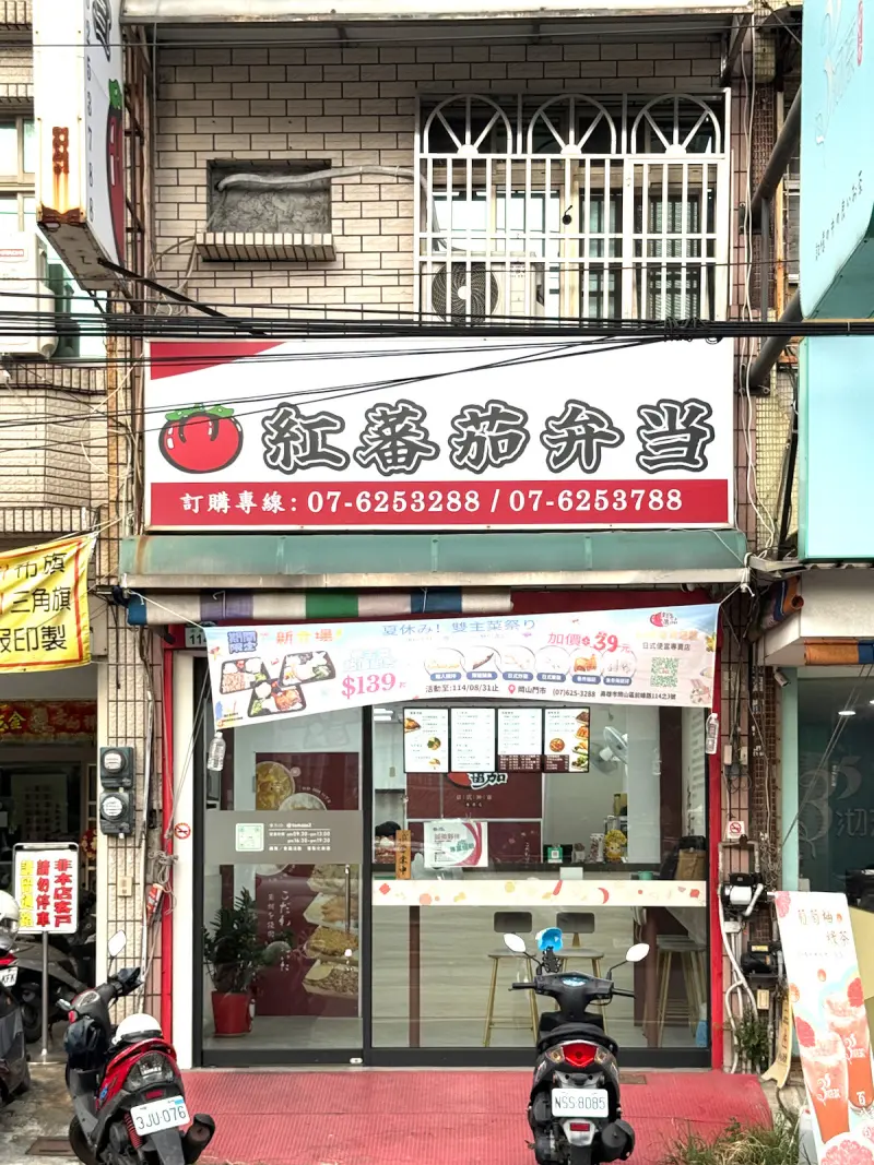 紅蕃茄日式便當專賣店的店面外觀，簡約明亮的兩層樓建築，搭配顯眼的紅色招牌與大片玻璃門，方便顧客一眼辨識。門口停著幾台機車，顯示出這家店的高人氣與外帶便利性。